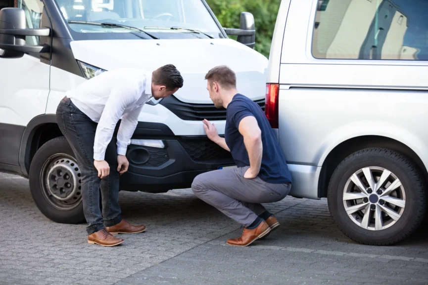 Two men inspecting front-end damage on a white car after a minor collision in a parking area.