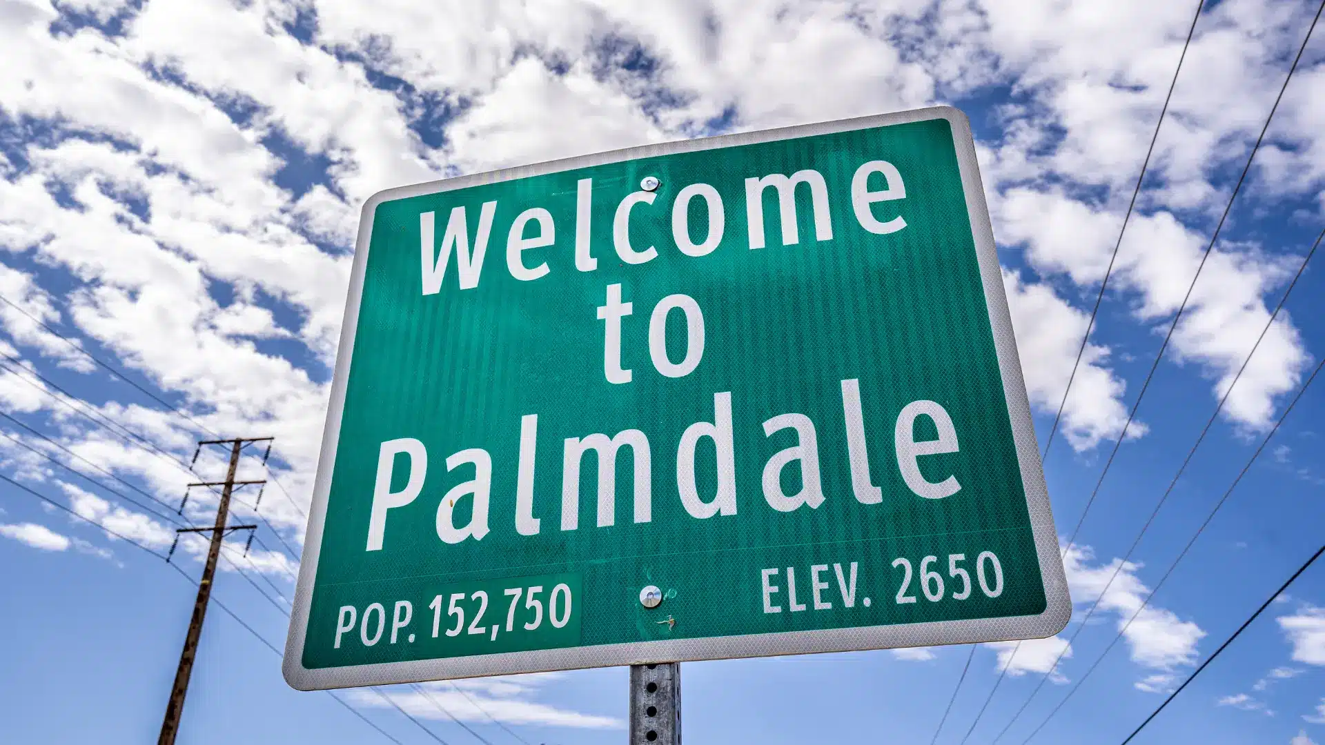 Green roadside sign reading “Welcome to Palmdale” against a partly cloudy blue sky.