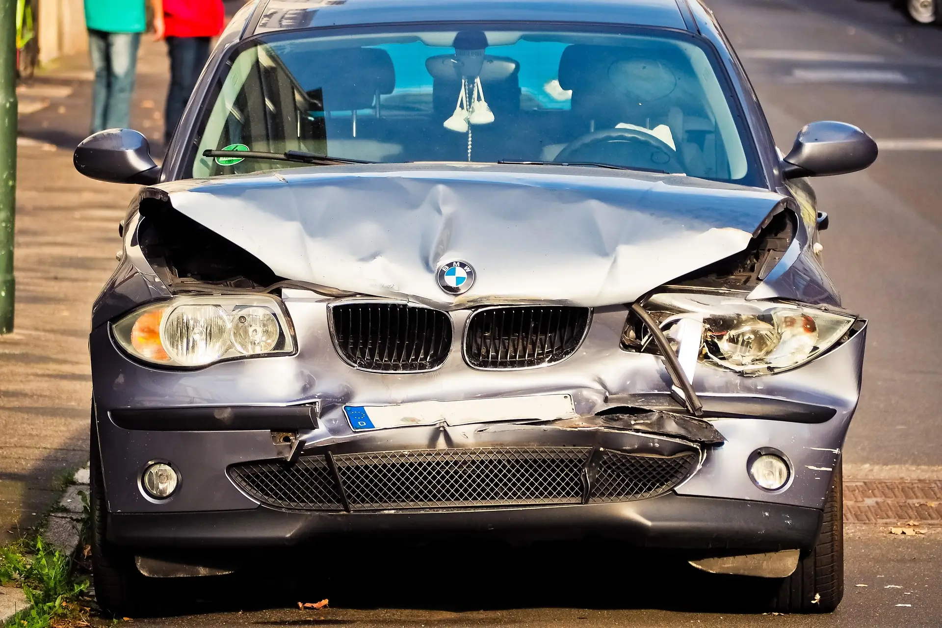A silver BMW car with severe front-end damage is parked on the side of a street.