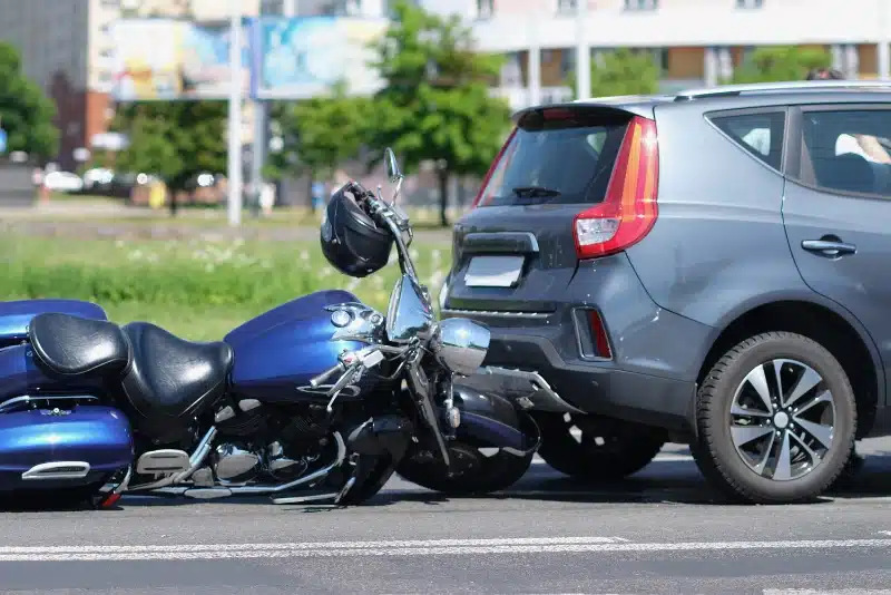 A blue motorcycle lies on its side after colliding with the rear of a gray car on a city street in daylight.