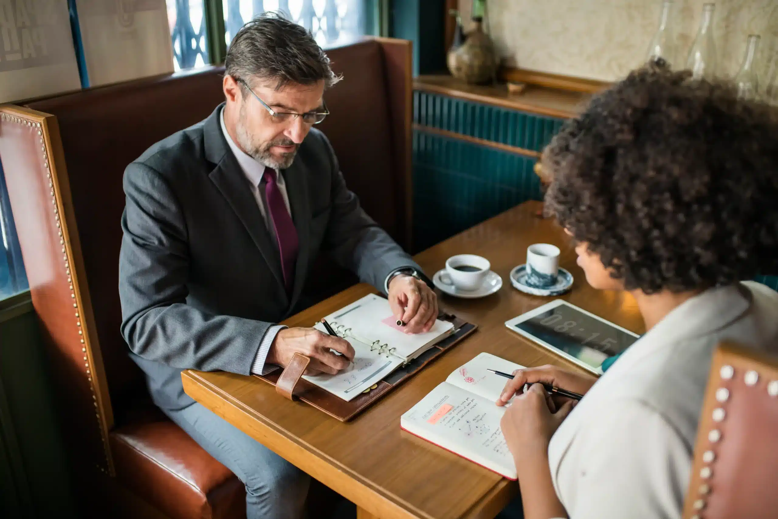 Two professionals sit across from each other at a café table, reviewing documents and taking notes during a business discussion.