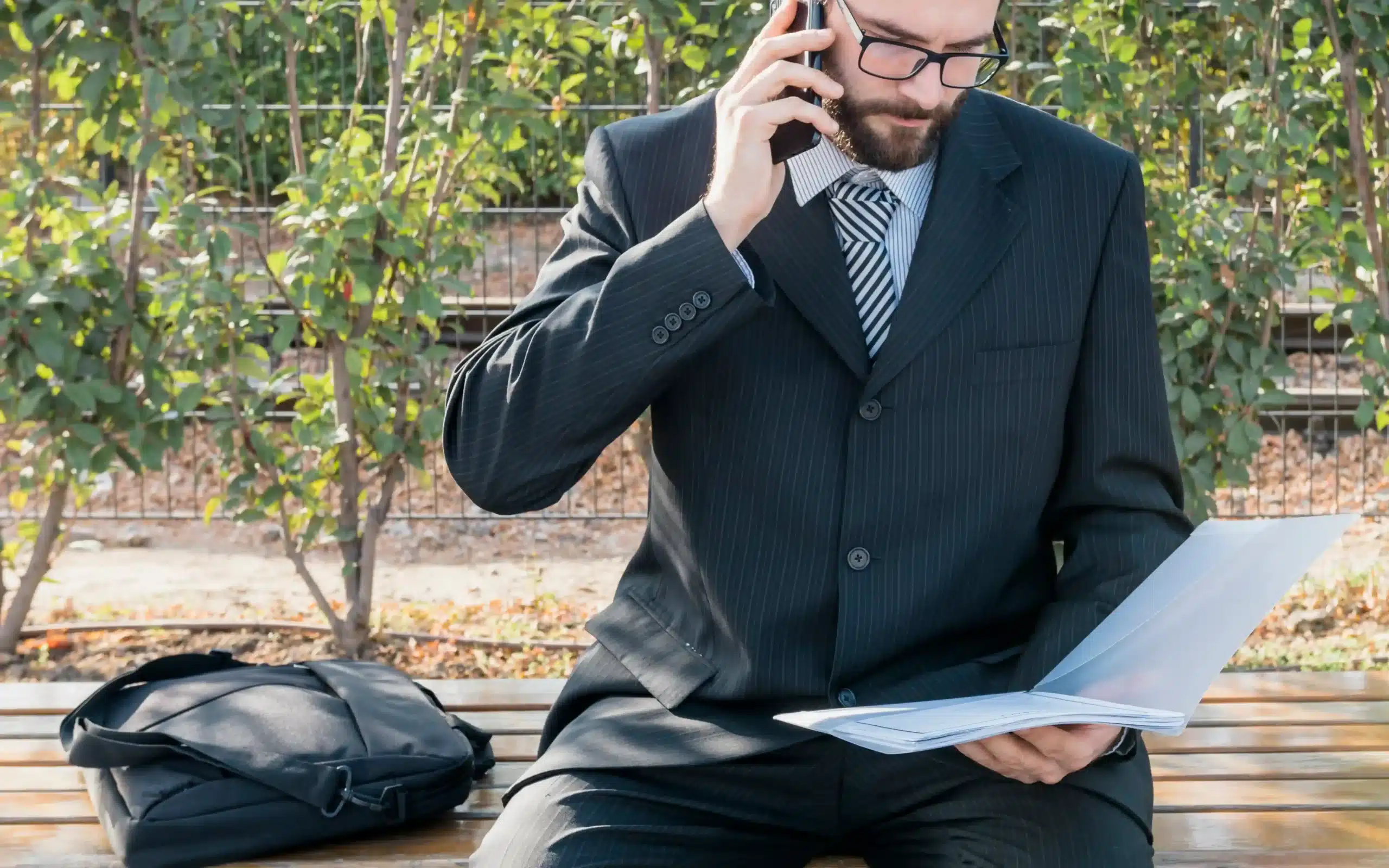 Business professional in a suit talking on the phone while reviewing documents on a park bench.