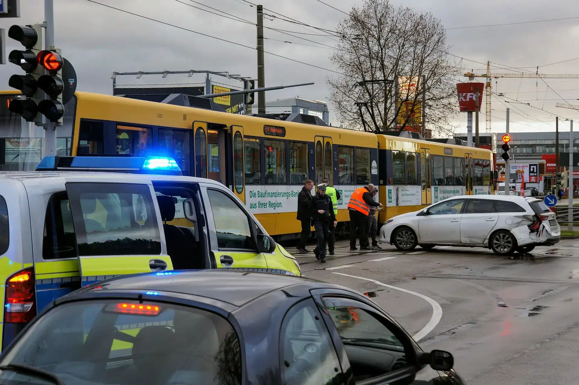 A yellow tram and a white car are involved in a collision at an intersection. Police vehicles with flashing lights are present, and officers assess the scene.