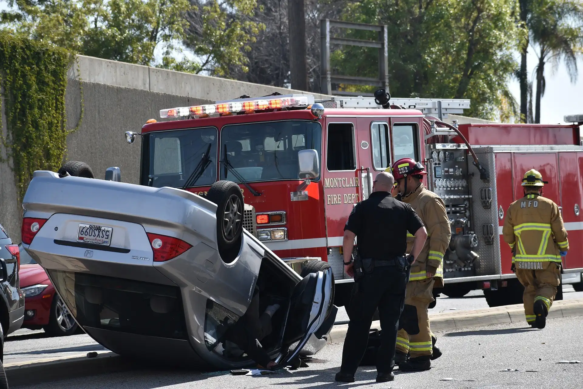 Overturned silver car on road with Montclair fire truck and responders. Police officer and firefighters assessing the scene. Bright daylight setting.