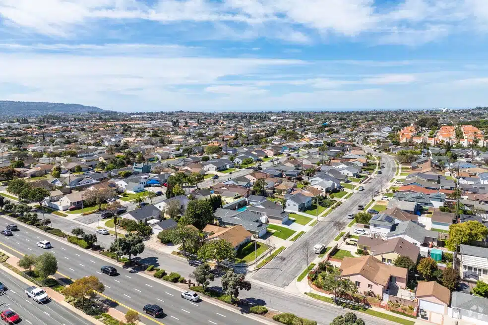 Aerial view of a suburban neighborhood, featuring rows of houses with similar architecture, manicured lawns, and a major road in the foreground under a partly cloudy sky.